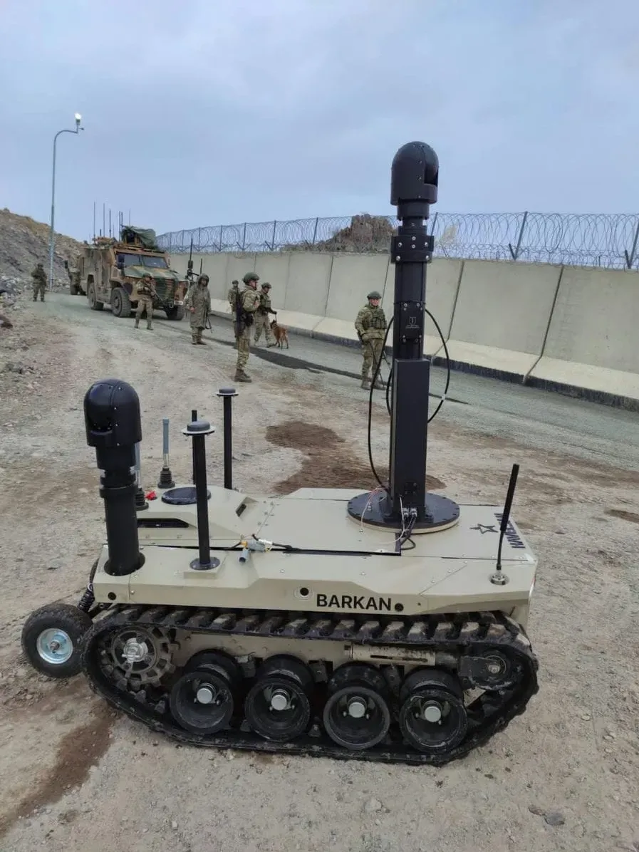 Çöl rengi BARKAN İKA üzerinde iki yükseltilmiş mast gimbali; arkada duvarın gerisinde askerler ve araçlar