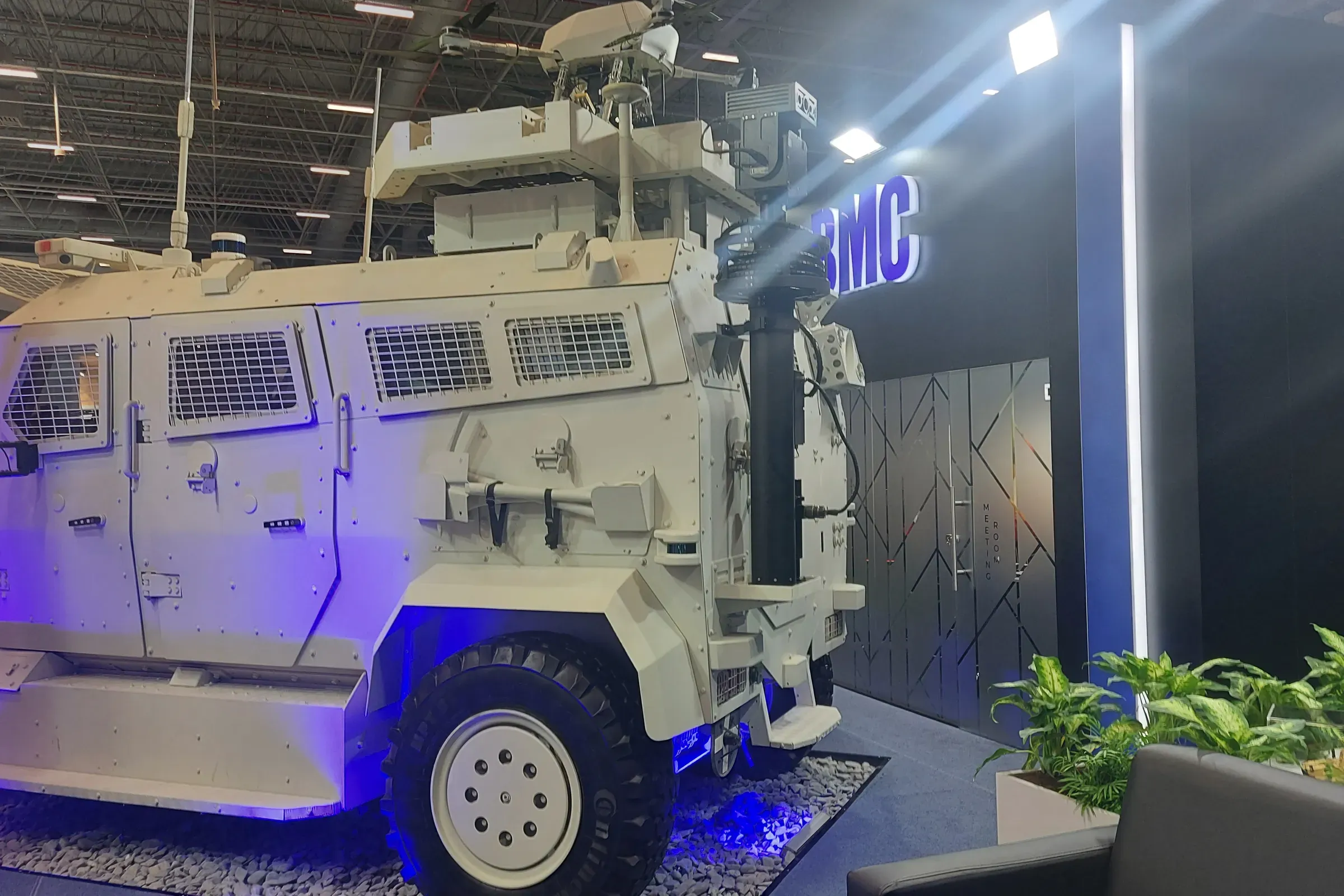 Stantta sergilenen beyaz zırhlı 4x4 araç; kabinin arkasında yükseltilmiş mast, mavi zemin aydınlatması