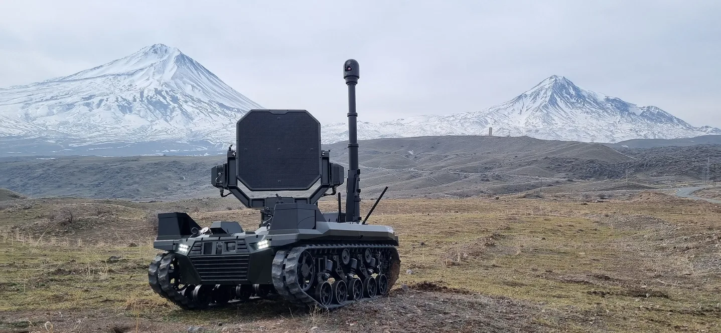 Karlı dağların önünde açık arazide duran paletli insansız kara aracı; üzerinde geniş akustik panel, arkasında yukarı çıkmış teleskopik mast ve ucunda stabilize gimbal