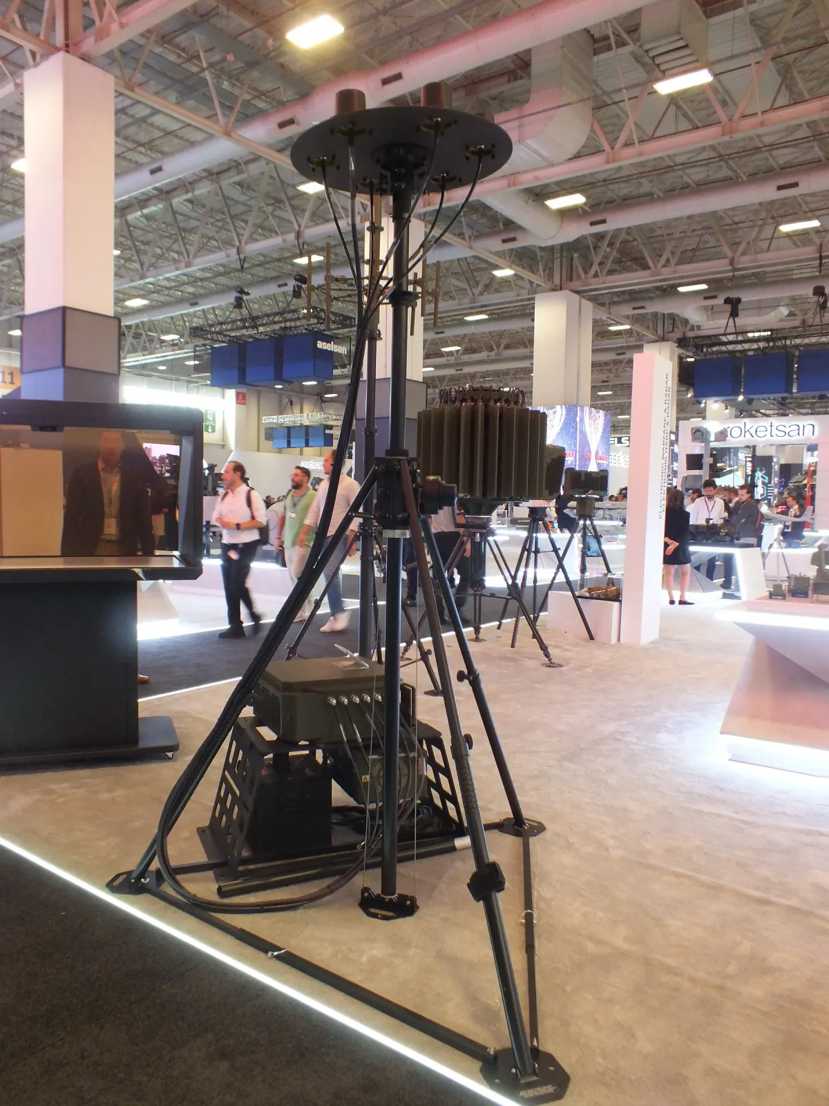 Fuar zemininde kurulmuş portatif tripod-mast; gergi halkaları ve faydalı yük başı görünüyor
