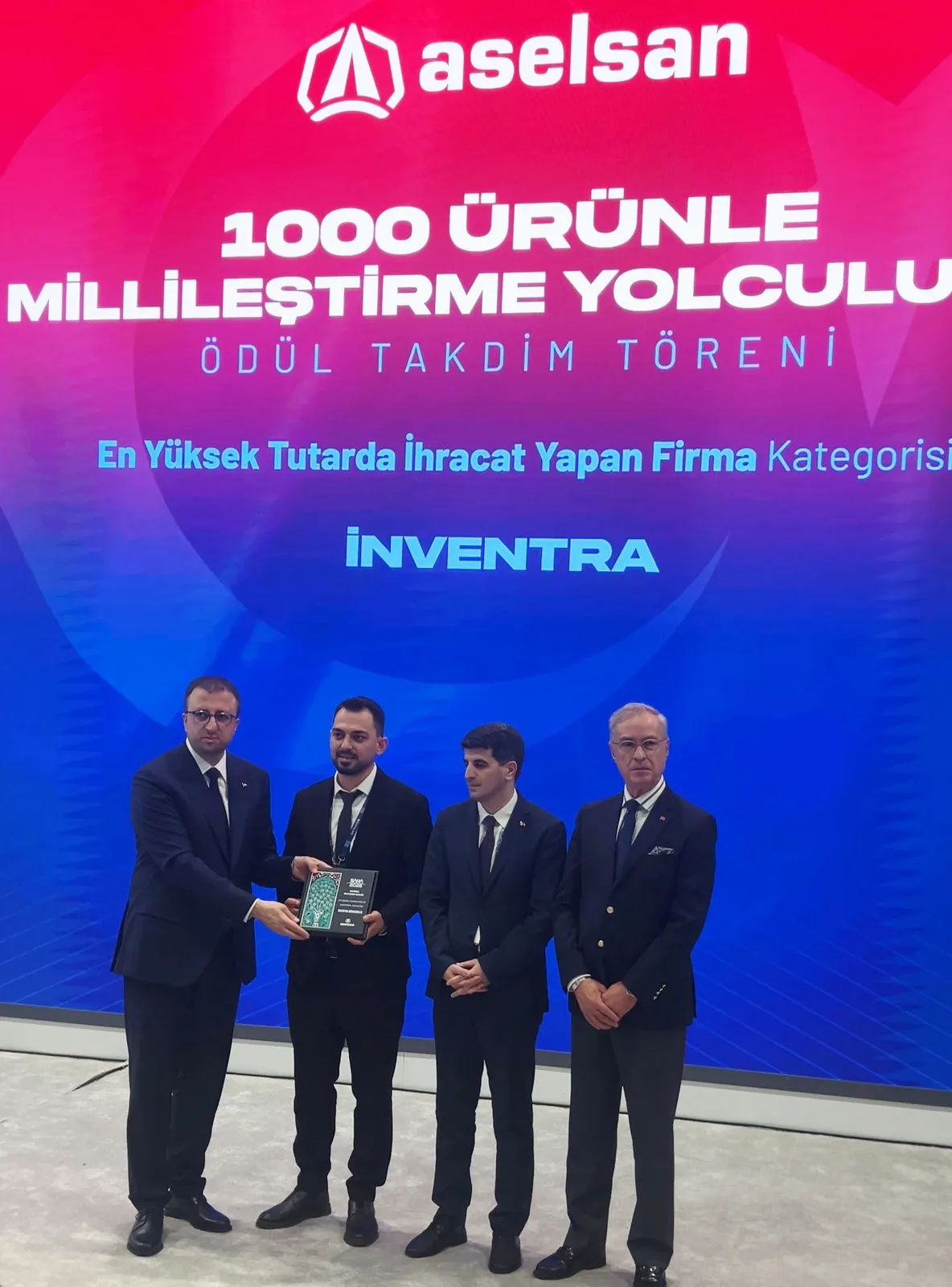 ASELSAN standında ödül takdimi: dört kişiden biri plaketi tutuyor; arkadaki ekranda “1000 Ürünle Millileştirme Yolculuğu — Ödül Takdim Töreni — En Yüksek Tutarda İhracat Yapan Firma Kategorisi — İnventra” yazıyor