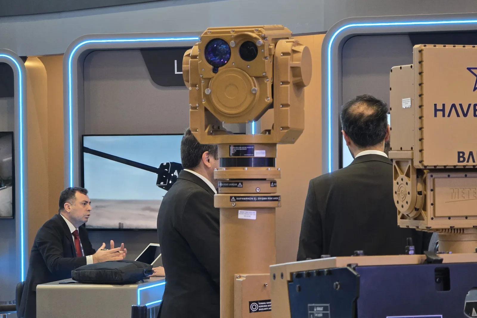 Önden görünen gimbal ve mastı; aracın çevresinde konuşan stand ziyaretçileri