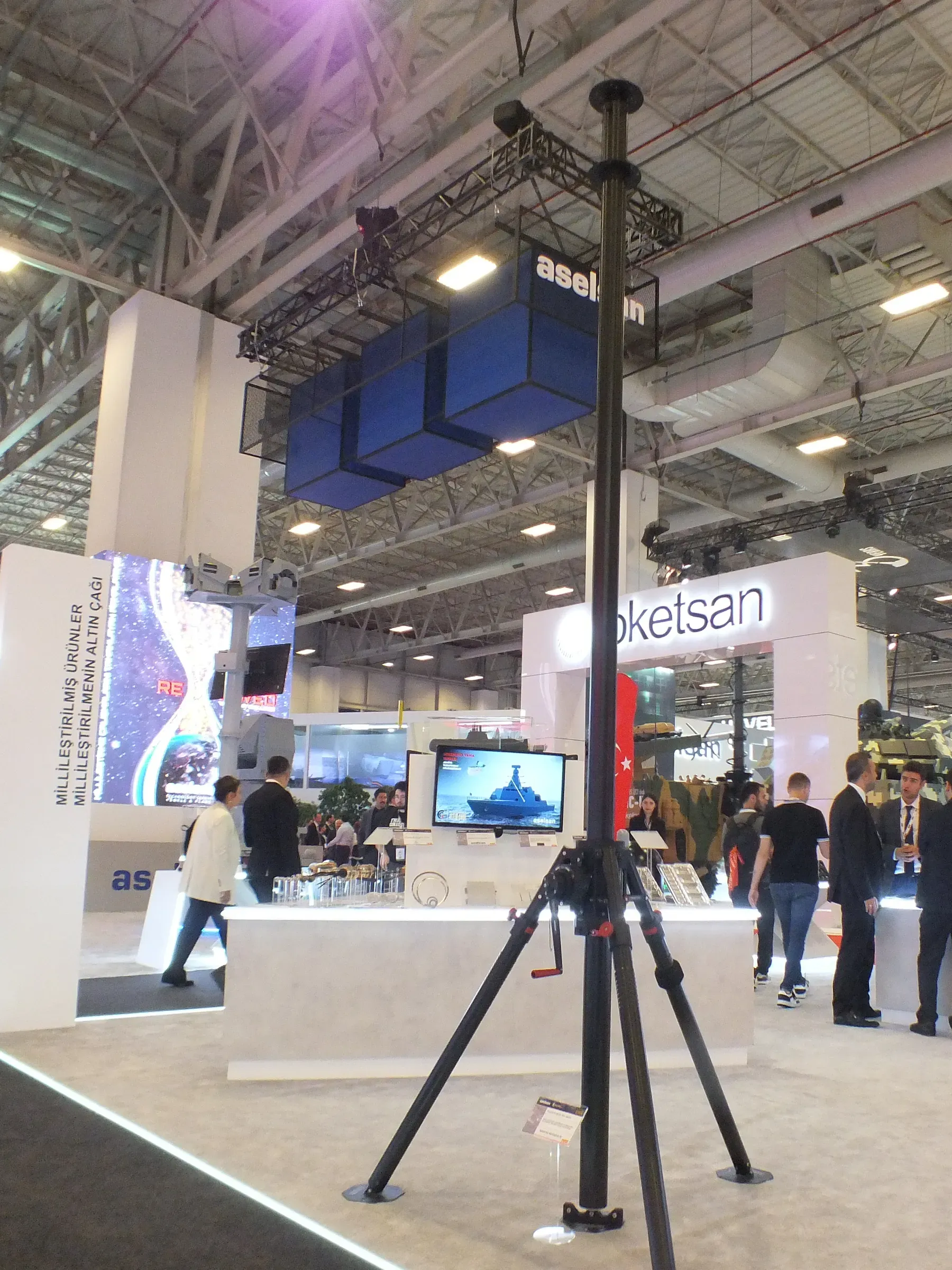 Fuar salonunda tripod üzerinde yükseltilmiş teleskopik mast; arkada Roketsan standı ve ziyaretçiler