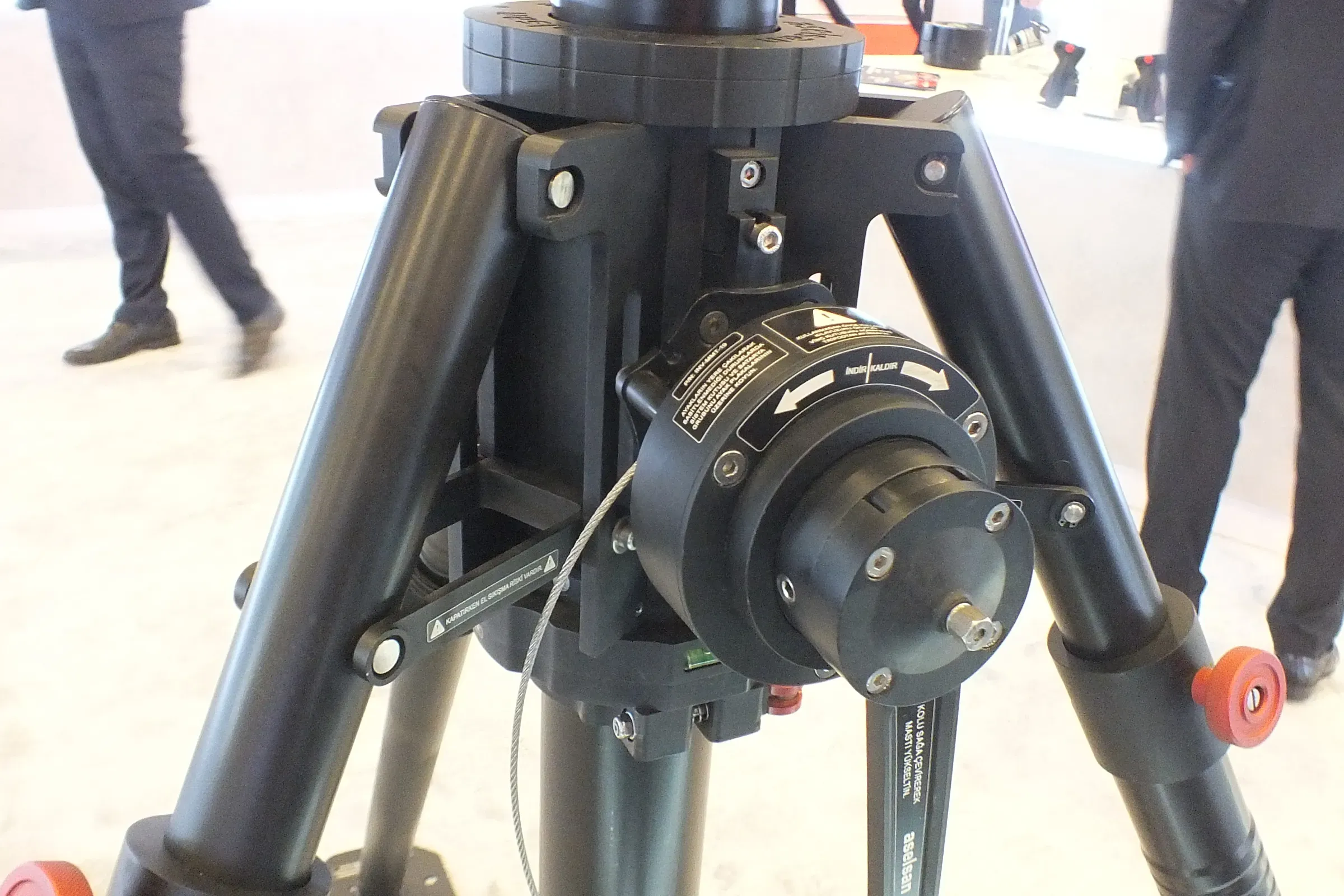 Kırmızı kelepçe topuzlarıyla tripod başı ve mast tabanının yakın çekimi; arkadan geçen ziyaretçiler