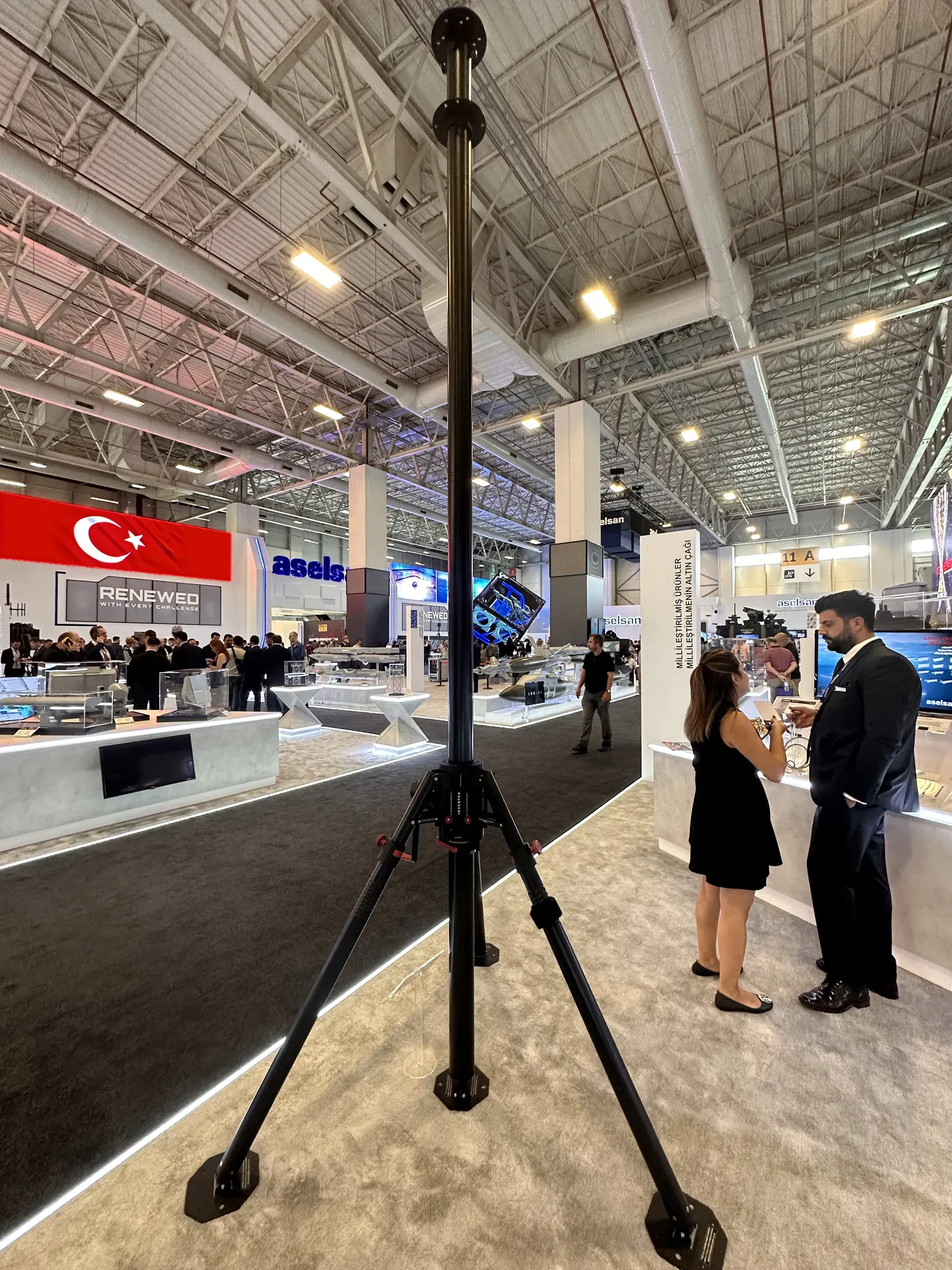 Stant halısında tripodu üzerinde açılmış teleskopik mast; yanında sohbet eden iki ziyaretçi