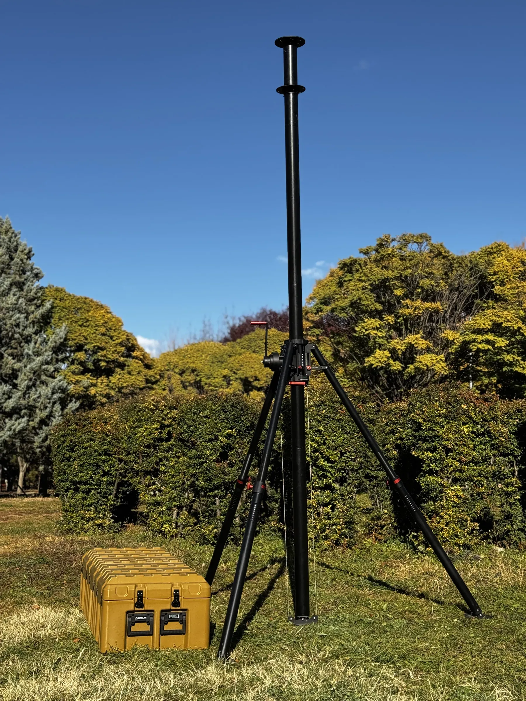 Çalıların önünde çimende duran açılmış tripod mast; dibinde zeytin yeşili taşıma sandığı