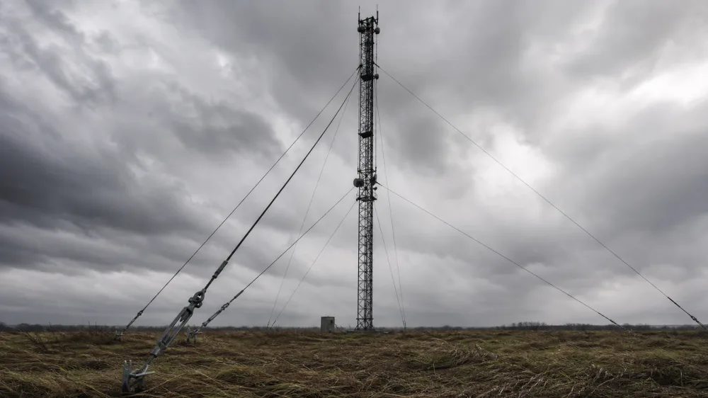 Ağır fırtına gökyüzüne karşı gergi halatlı kafes mast; çapraz gergi halatları gerdirici ve zemin ankrajlarıyla tutuluyor, altındaki ot rüzgârla yatmış